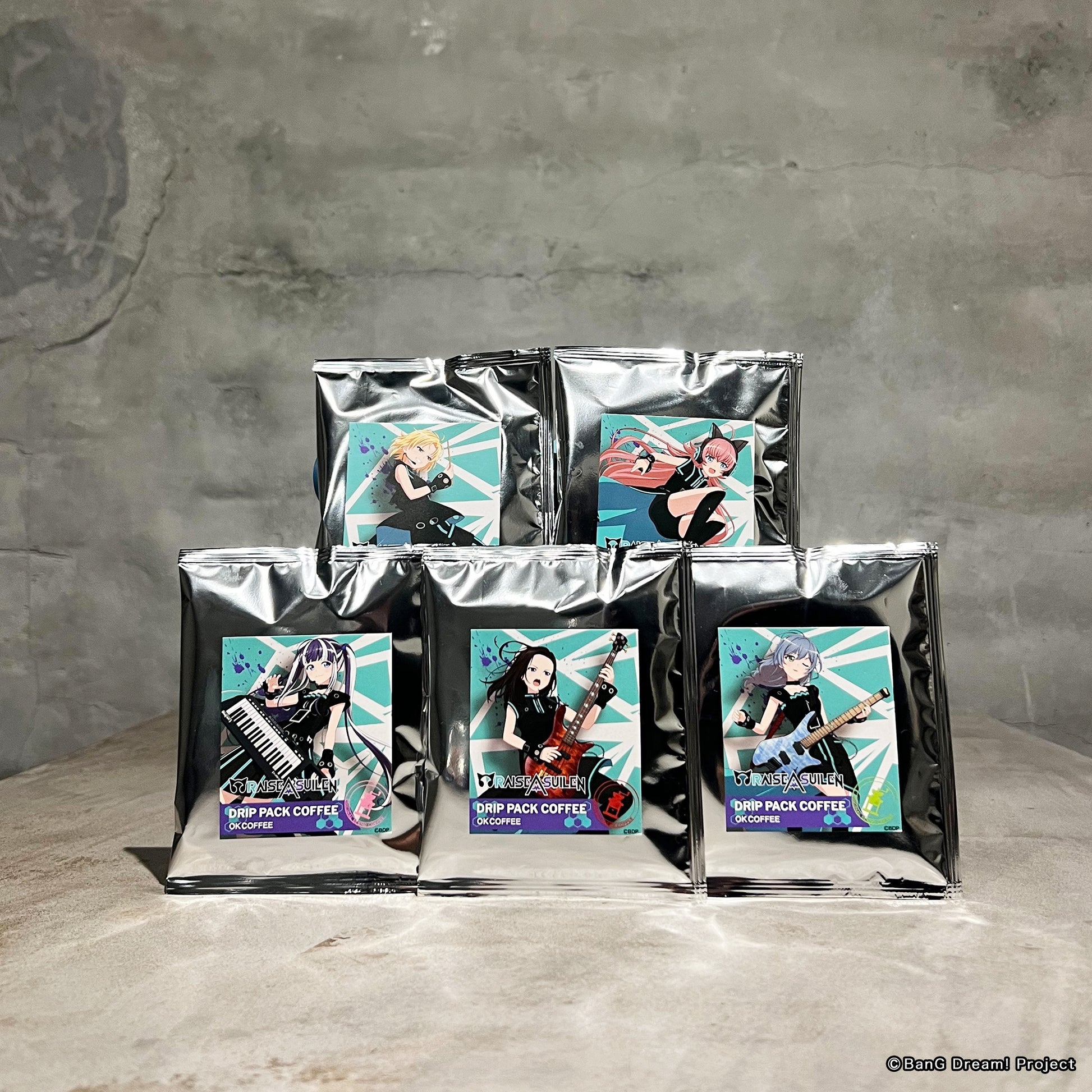 Five sealed silver drip-pack coffee pouches with RAISE A SUILEN member illustration labels arranged on a tabletop.