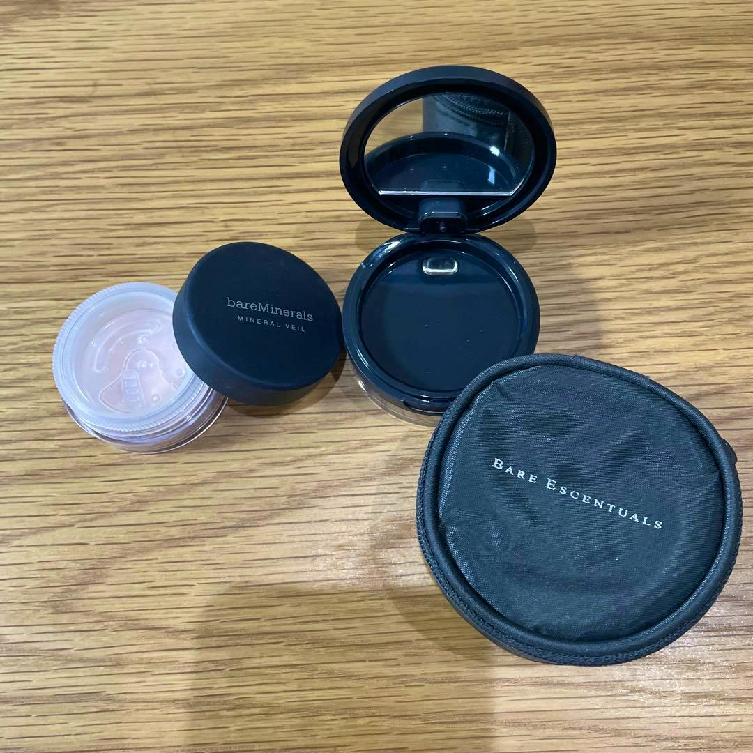 A bareMinerals Mineral Veil refill jar, an open black refill compact with mirror, and a round Bare Escentuals zip pouch on a wooden surface.