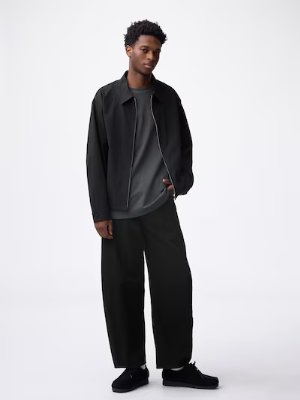 A model wearing black barrel-leg men's jeans with a jacket and sneakers on a white background.