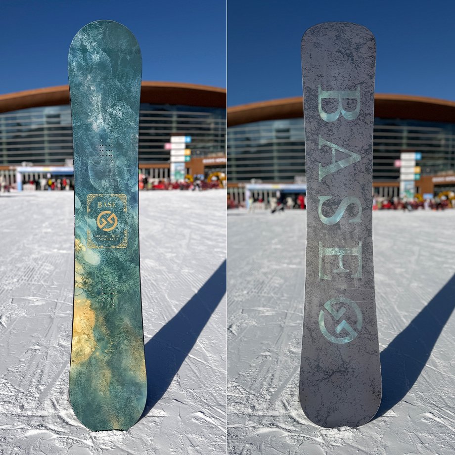 A snowboard photographed upright on snow showing the board's top graphic and base design with a building in the background.