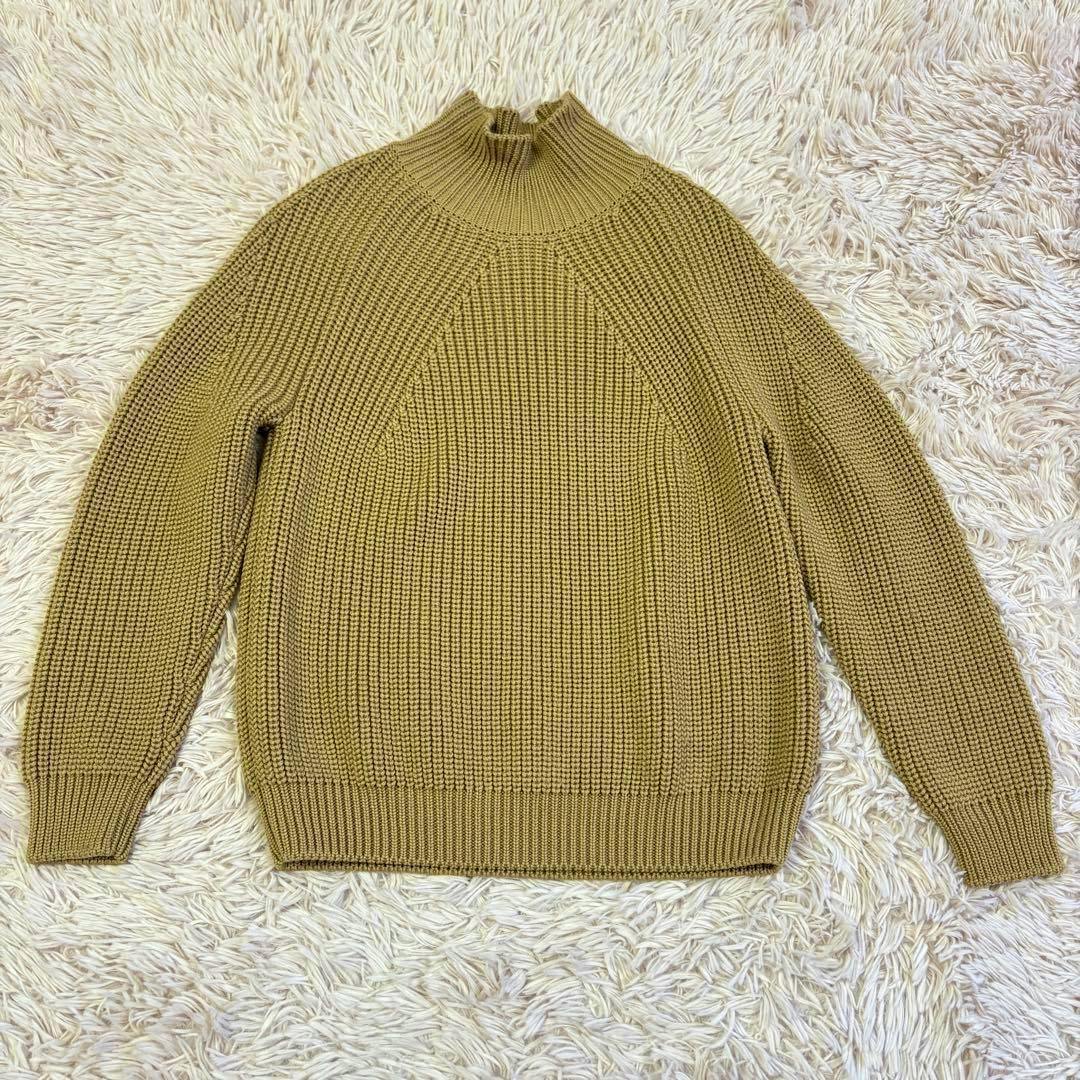 Camel-colored BATONER wool sweater laid flat on a light shag rug, front view showing ribbed knit and mock neck.