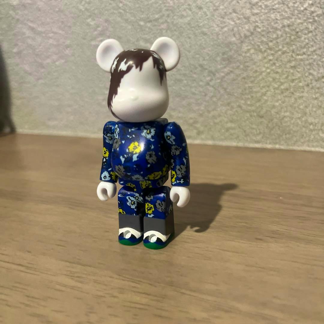 Front view of a BE@RBRICK Raf Simons figure with a white head painted brown at the top and a blue floral-patterned body standing on a wooden surface.