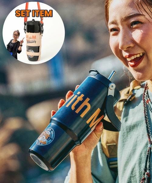 A person holding a dark blue stainless steel bottle with visible "with M" lettering and a flip-style lid outdoors.