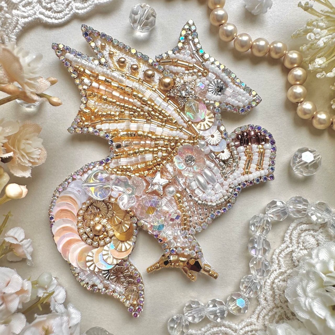 A beaded brooch shaped like a winged animal, decorated with sequins, crystals, and pearls on a lace background.