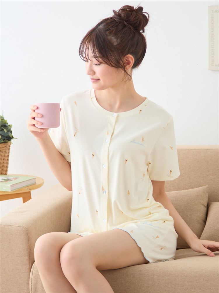 A woman models a cream short-sleeve patterned pajama top and matching shorts while seated on a sofa holding a pink mug.