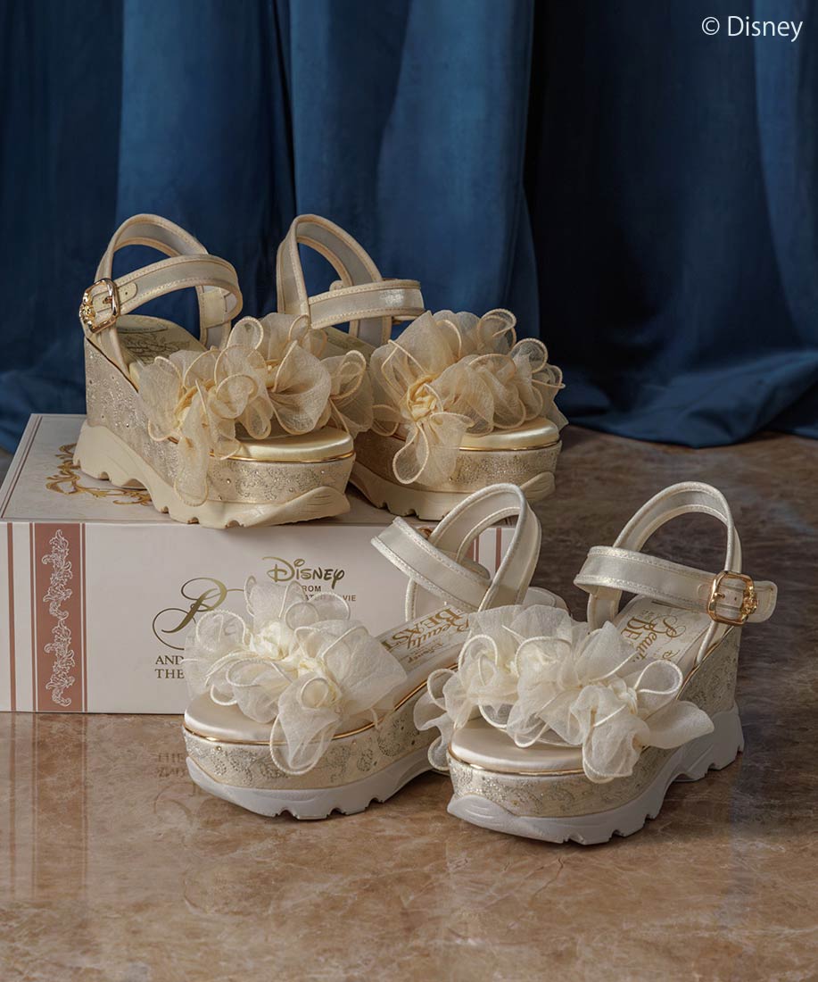 Two pairs of synthetic leather platform dress-up sports sandals with ruffled organza flower decorations displayed on a floor with a decorative box.