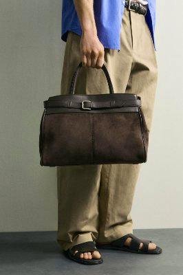 A person is holding a brown belt-detail combination handbag by its top handles.