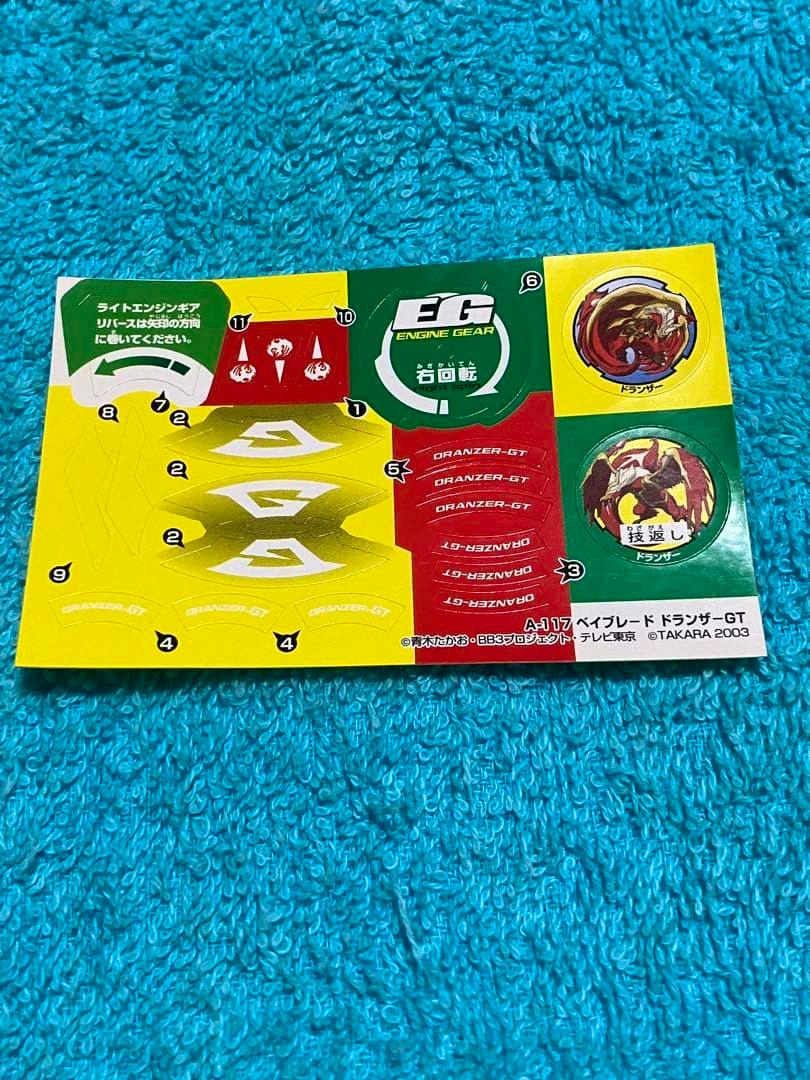 A sheet of Beyblade A-117 Dranzerzer GT paper stickers laid on a blue textured towel showing multiple colorful sticker designs and printed text.