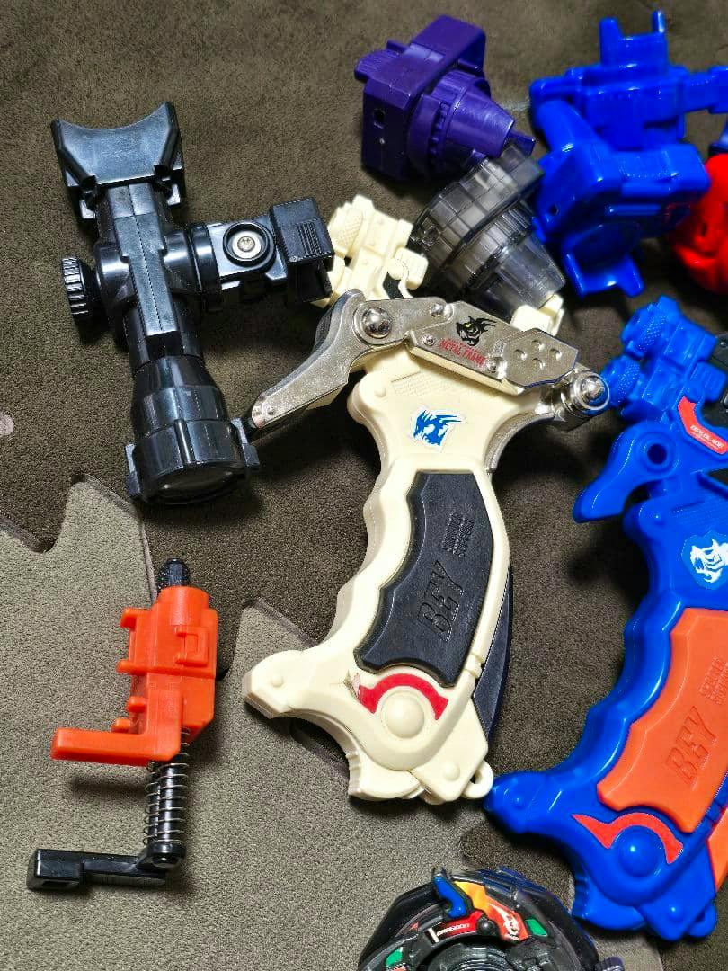 Close-up photo of Beyblade customization grips/shooters (cream and blue) alongside launcher components and a partial Beyblade top.
