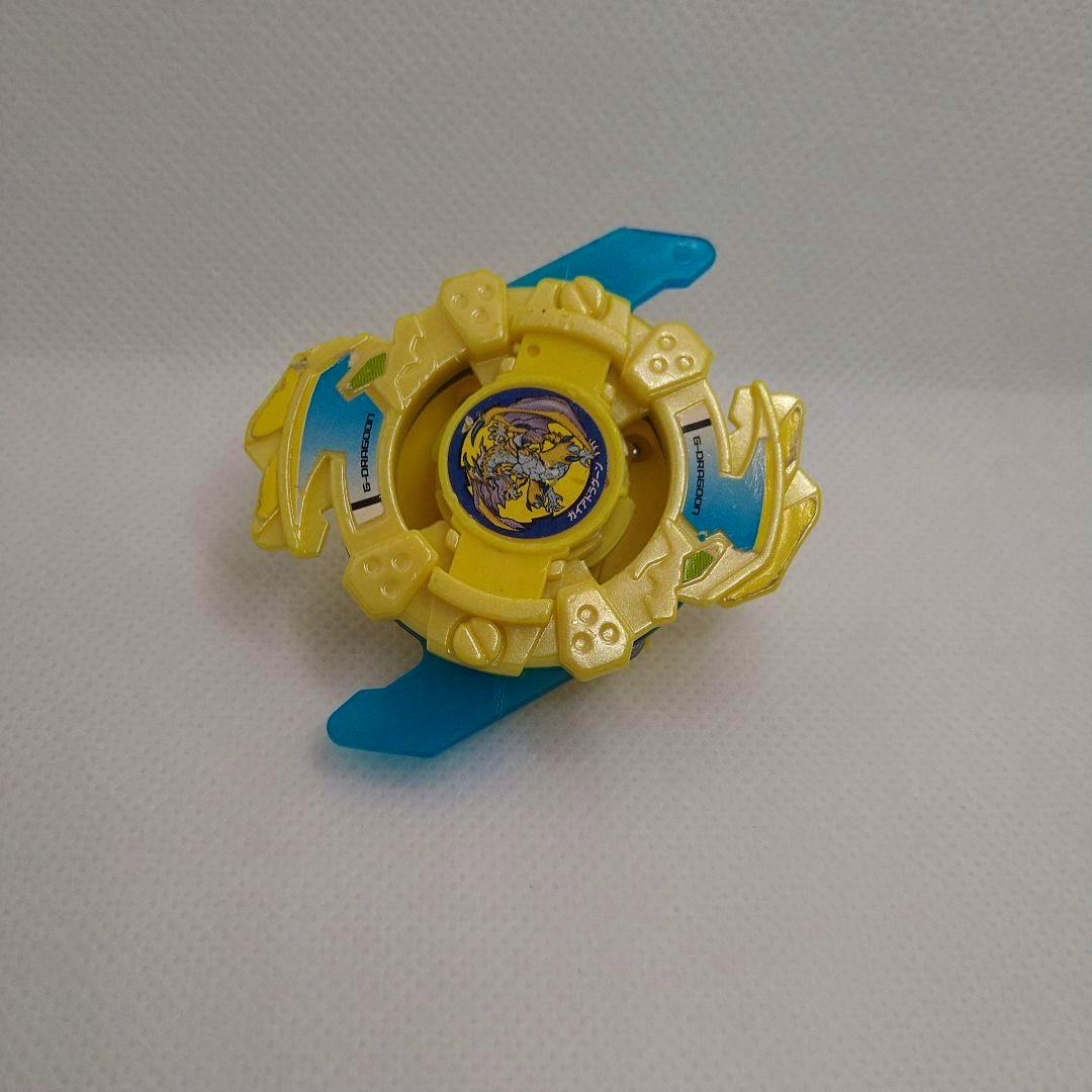 Top view of the yellow Gaia Dragoon Beyblade toy (黄龍 Ver.) showing yellow plastic parts, blue blades, and a central sticker artwork.