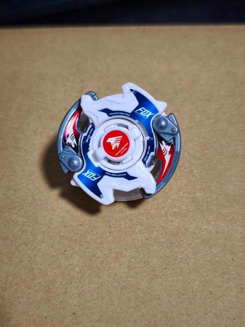 Top view of the Beyblade HMS Phantom Fox toy showing white and blue plastic layers and a red red center on a brown surface.