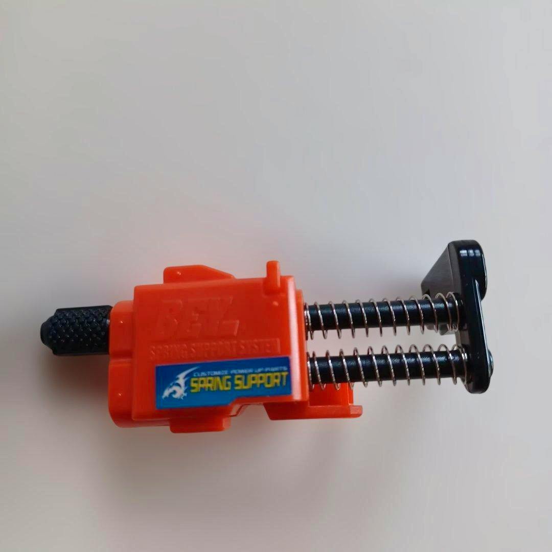Orange plastic Beyblade spring support toy part with two metal springs and a black adjustment knob on a white background.