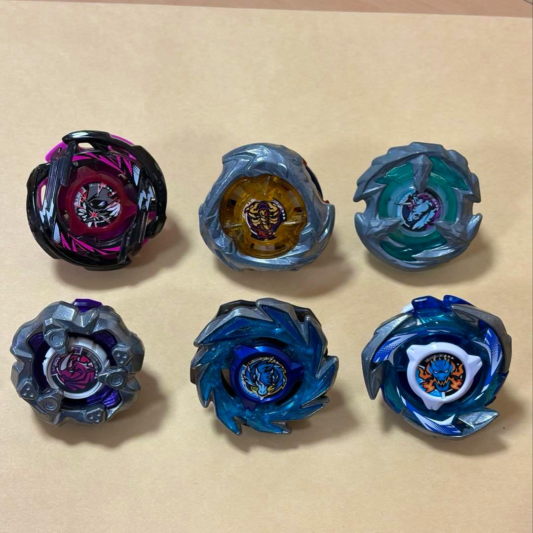 Six Beyblade X toy tops arranged in two rows on a beige surface, showing different colors and face stickers.