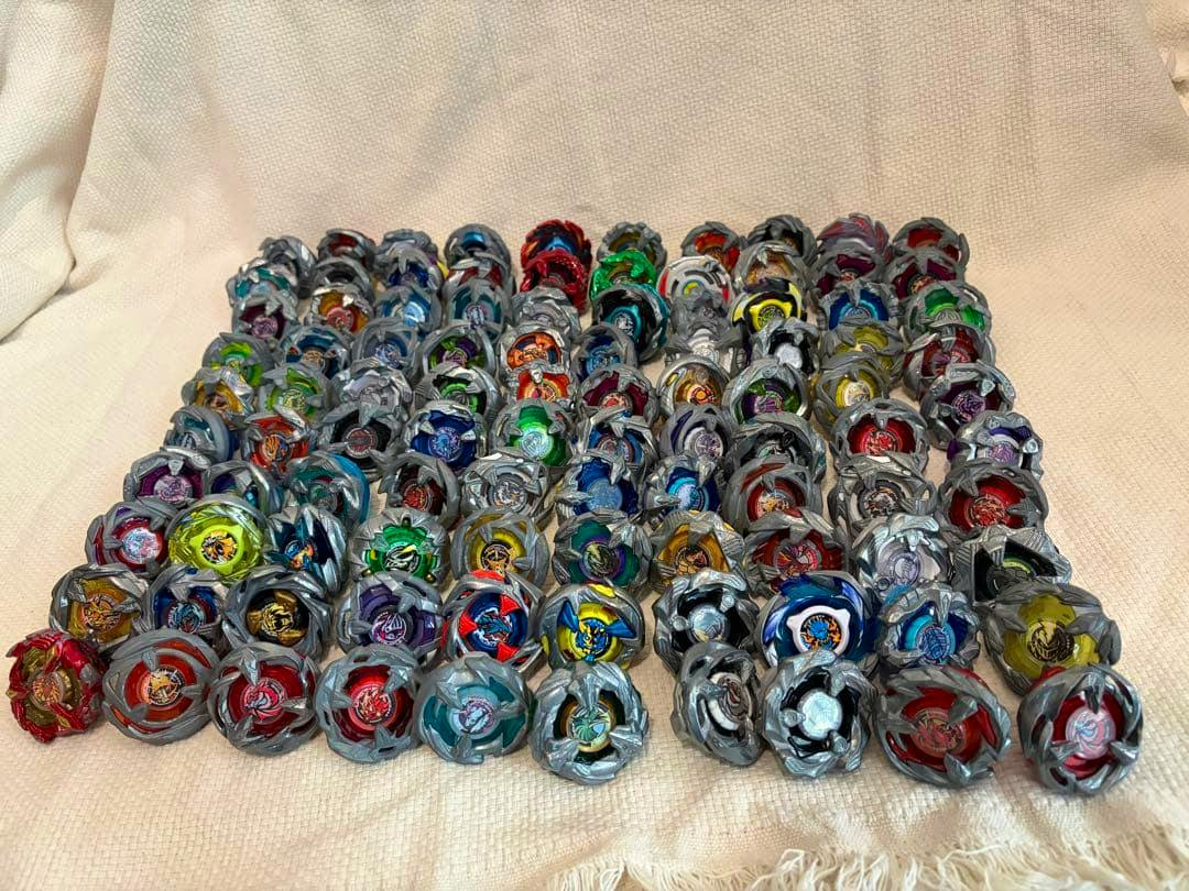 Many Beyblade X spinning tops arranged in rows on a light-colored fabric background.