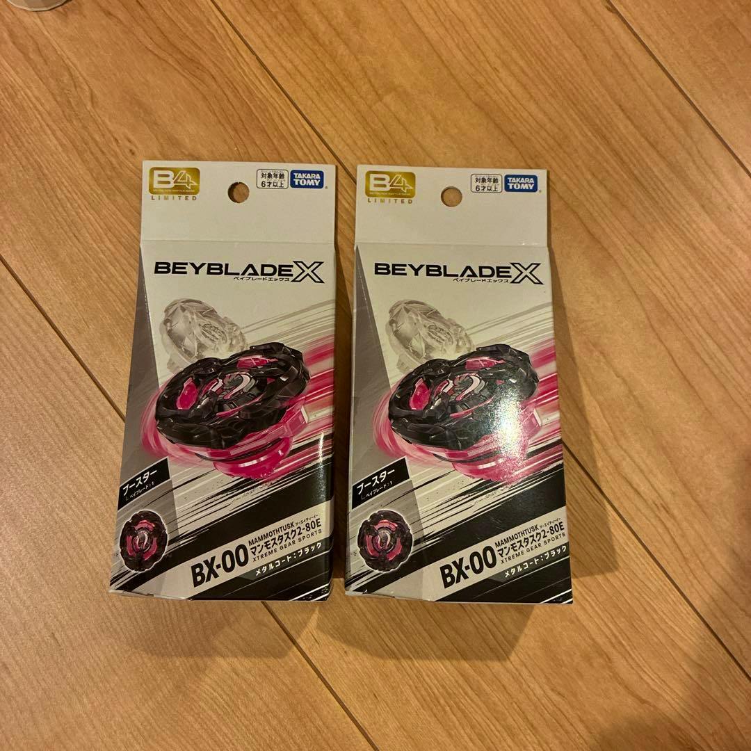 Two unopened Beyblade X BX-00 Mammoth Tusk (2-80E) toy packages shown front-facing on a wooden floor.