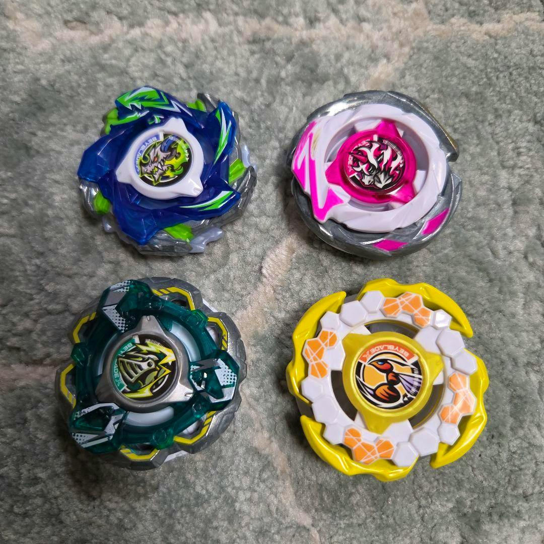 Four assembled Beyblade X CX parts in blue, pink, green, and yellow displayed on a carpeted surface.