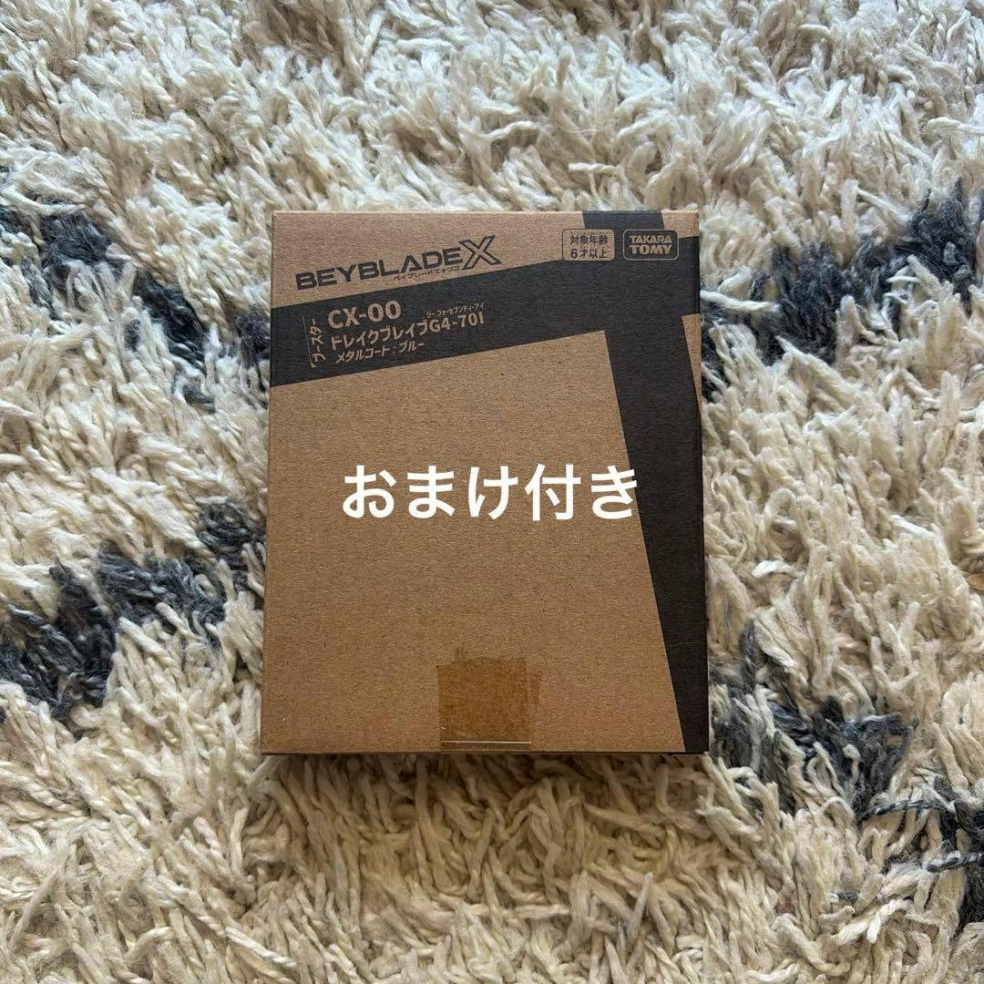 Cardboard Beyblade X product box labeled CX-00 ドレイクブレイブG4-70I メタルコート:ブルー on a rug.