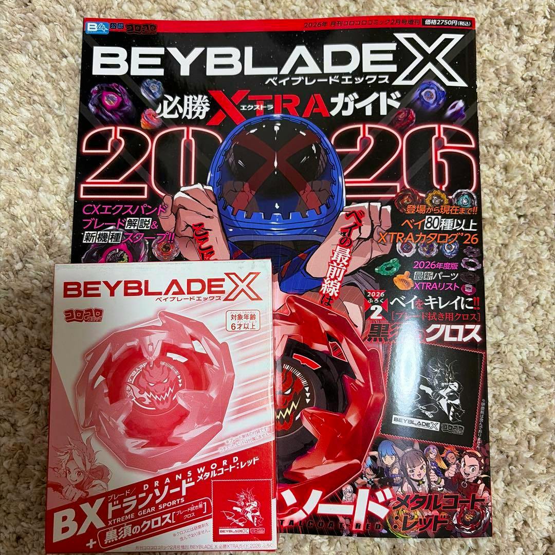 Photograph of the front cover of the BEYBLADE X 必勝XTRAガイド 2026 book lying on a carpet with a BEYBLADE X product box partially overlapping the lower left of the cover.