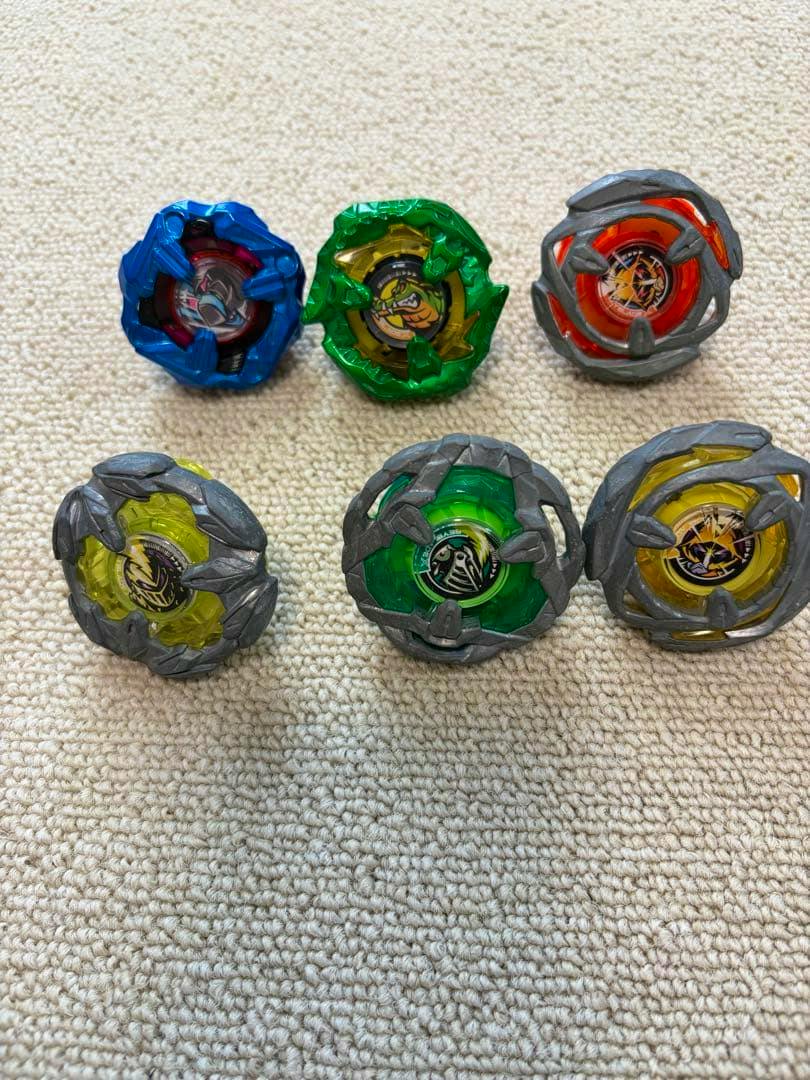 Six Beyblade X spinning tops arranged on a carpet showing their top faces.