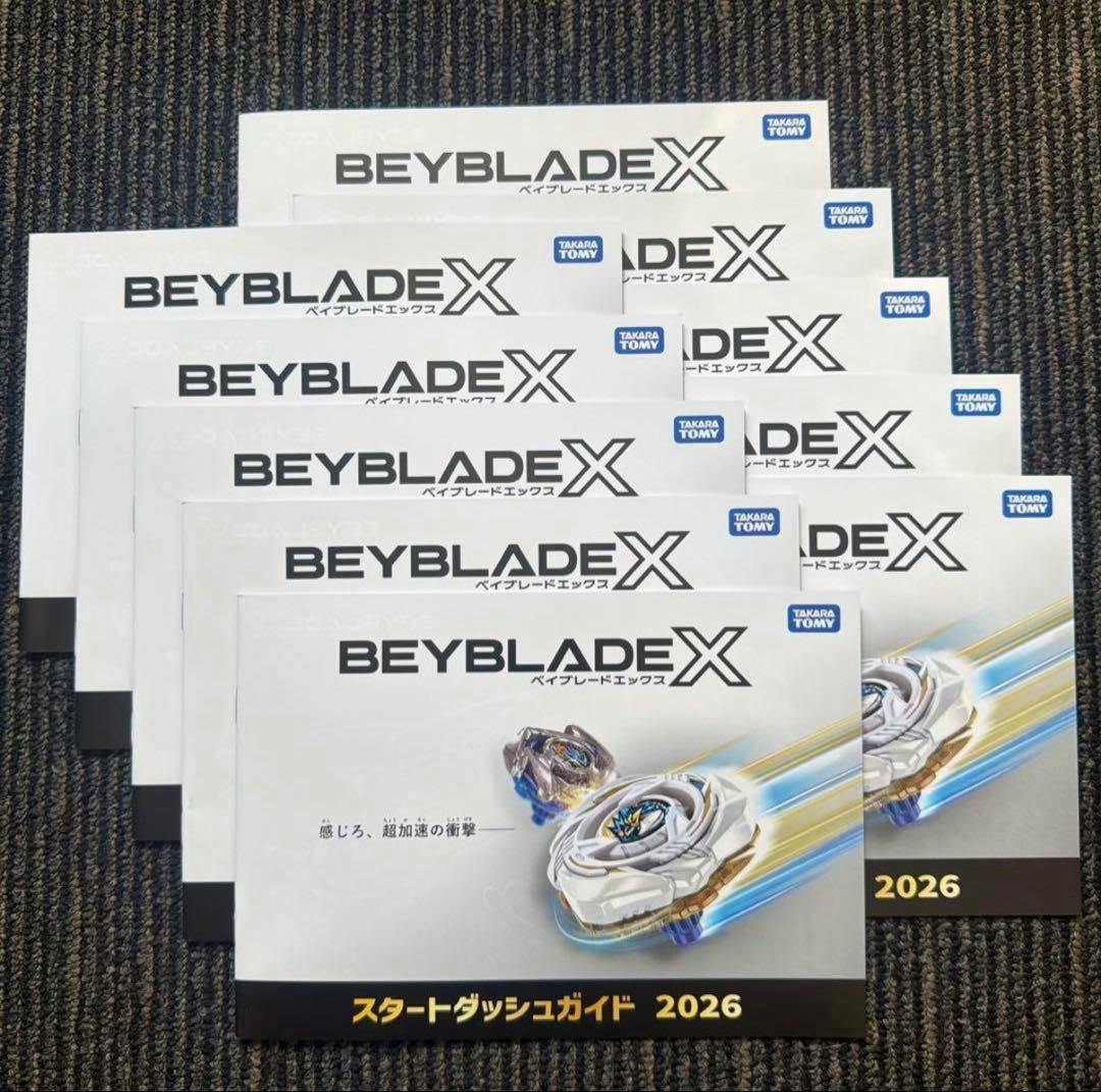 Photograph of multiple BEYBLADE X スタートダッシュガイド 2026 pamphlets arranged overlapping on a carpeted surface showing the cover and title.