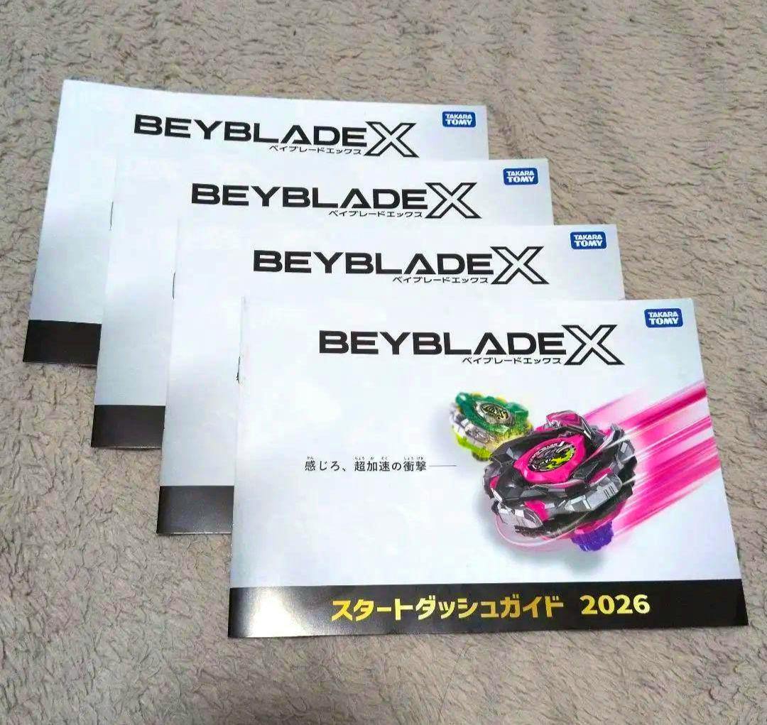 Four copies of the Beyblade X "Start Dash Guide 2026" booklets arranged overlapping on a textured (carpeted) surface.