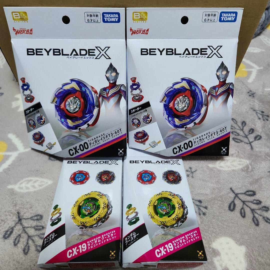 Four boxed Beyblade X products, two CX-00 Tigarage boxes and two CX-19 random booster boxes, arranged on a patterned surface.