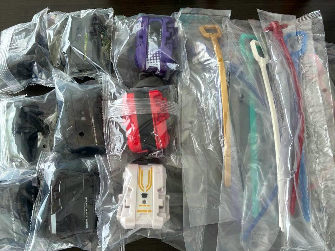 Multiple packaged Beyblade-style launcher bodies and several plastic winder/launcher handles in individual clear bags laid out on a dark surface.