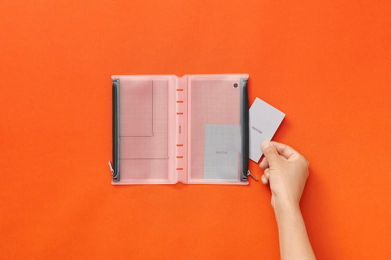 Open transparent zipper case (bible size) with two zippered pockets shown flat on an orange background, a hand holding a card at the right.