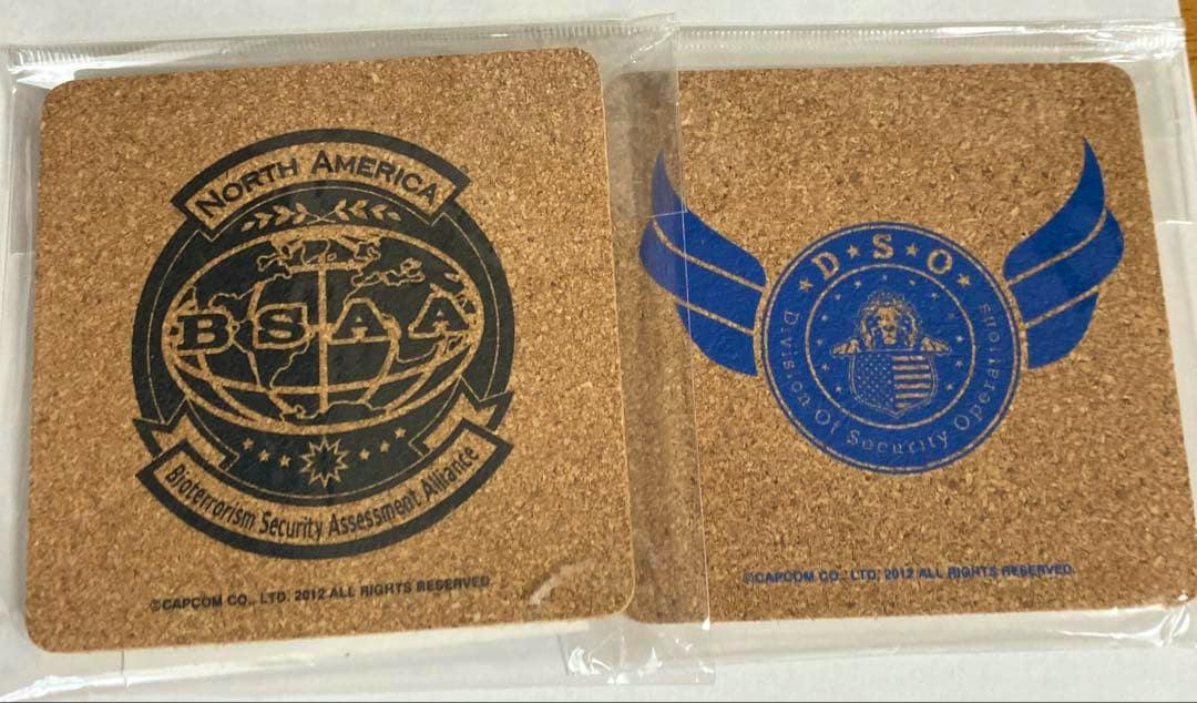 Two cork coasters side by side in plastic packaging, one printed with a BSAA logo and the other with a DSO winged emblem.