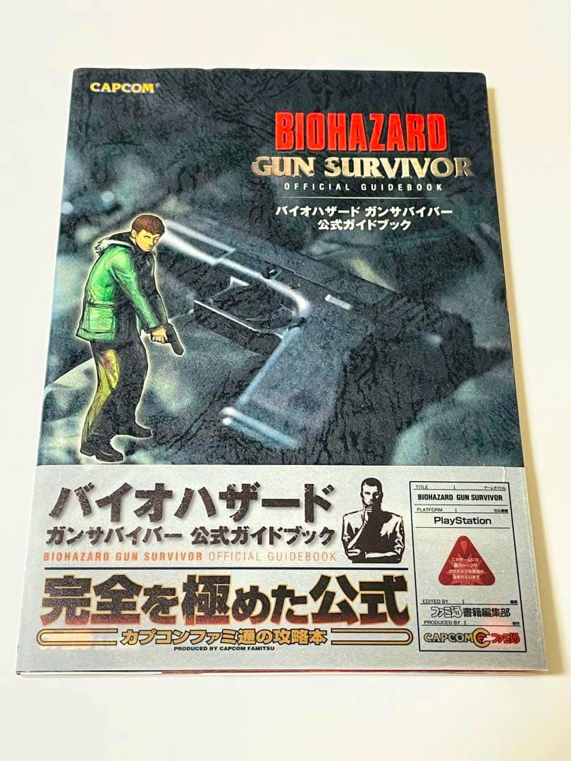 Front cover of the Biohazard Gun Survivor official guidebook showing cover art of a character with a handgun and a silver obi band at the bottom.