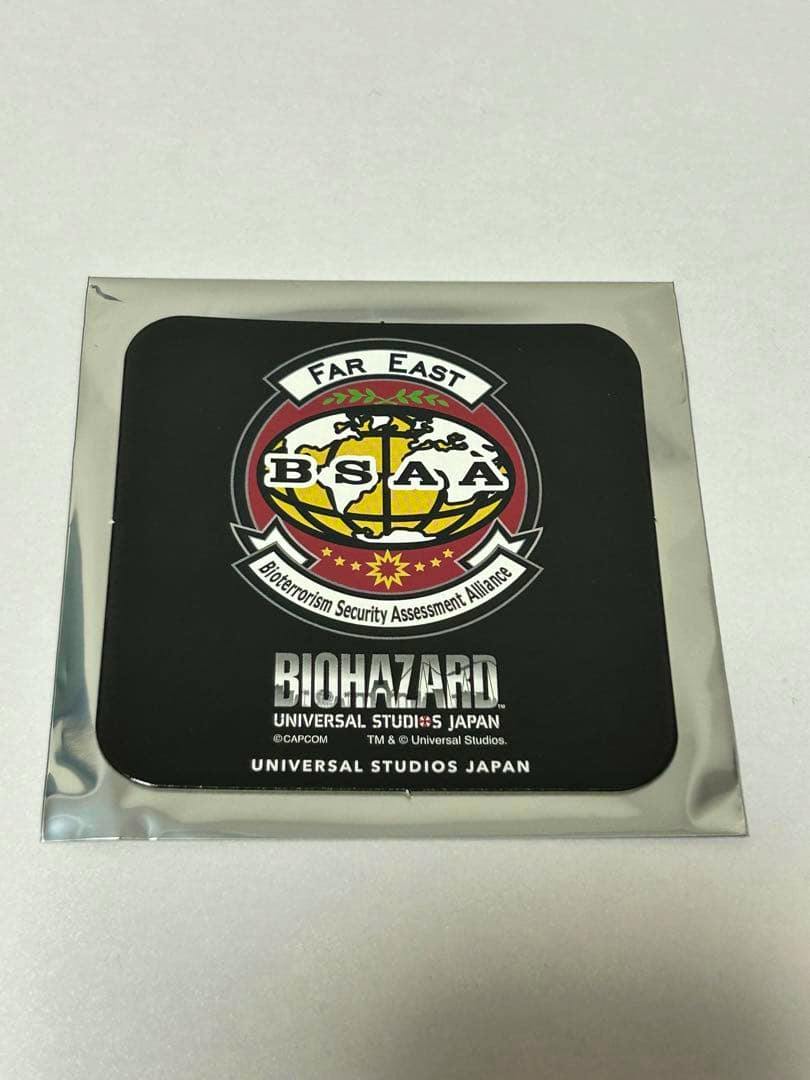 A black square paper coaster in silver packaging showing the BSAA emblem and the text "BIOHAZARD UNIVERSAL STUDIOS JAPAN."