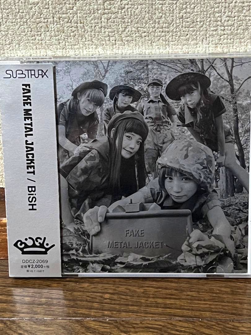 Front cover of the BISH 'FAKE METAL JACKET' CD showing a black-and-white photo of the group and a metal box labeled 'FAKE METAL JACKET' with an obi strip at left.