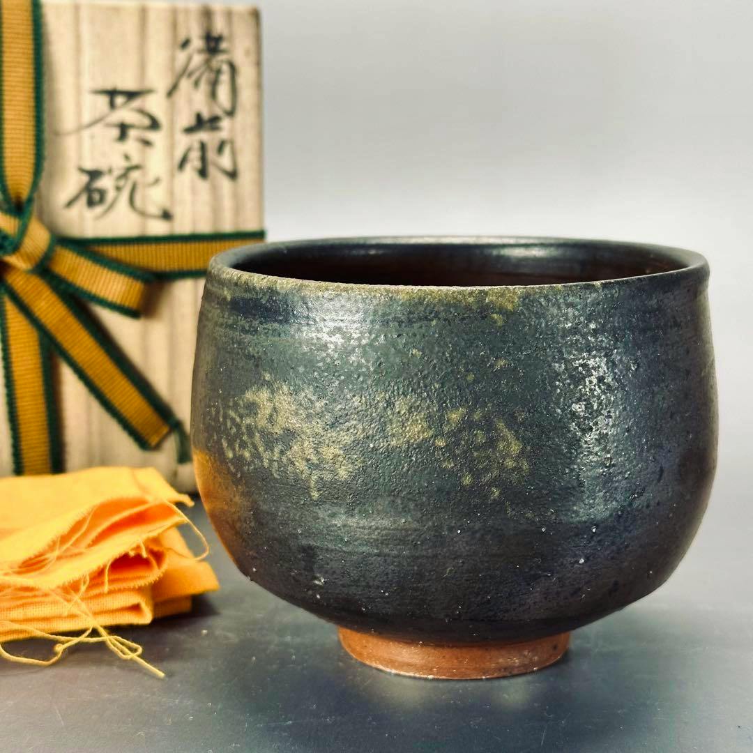 Front view of the ceramic bowl showing its dark glazed surface, with the wooden storage box and yellow cloth visible behind it.
