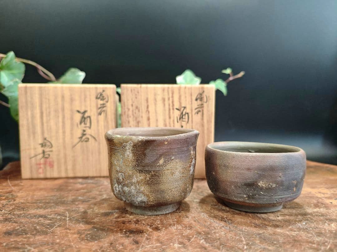 Two Bizen-yaki sake cups (guinomi) displayed on a wooden surface with their signed wooden storage boxes behind them.