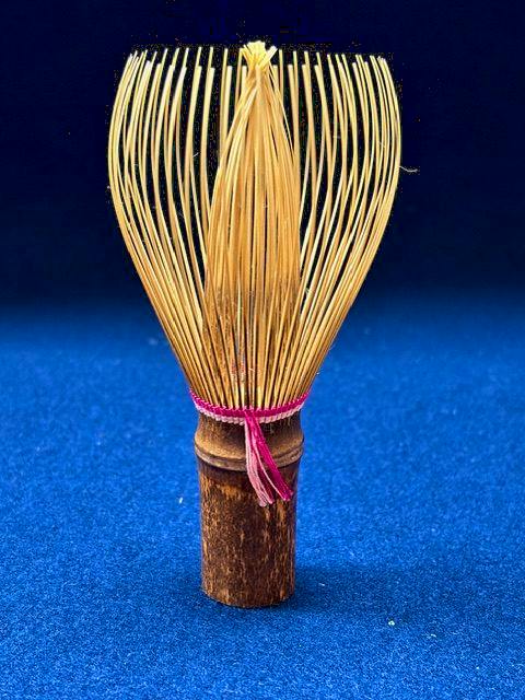 A bamboo tea whisk (chasen) with pink-colored binding displayed upright on a blue background.