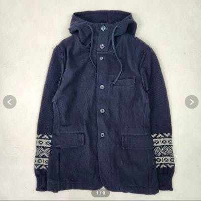 Black Comme des Garçons men's wool jacket laid flat showing hood, button front, and patterned sleeve cuffs.