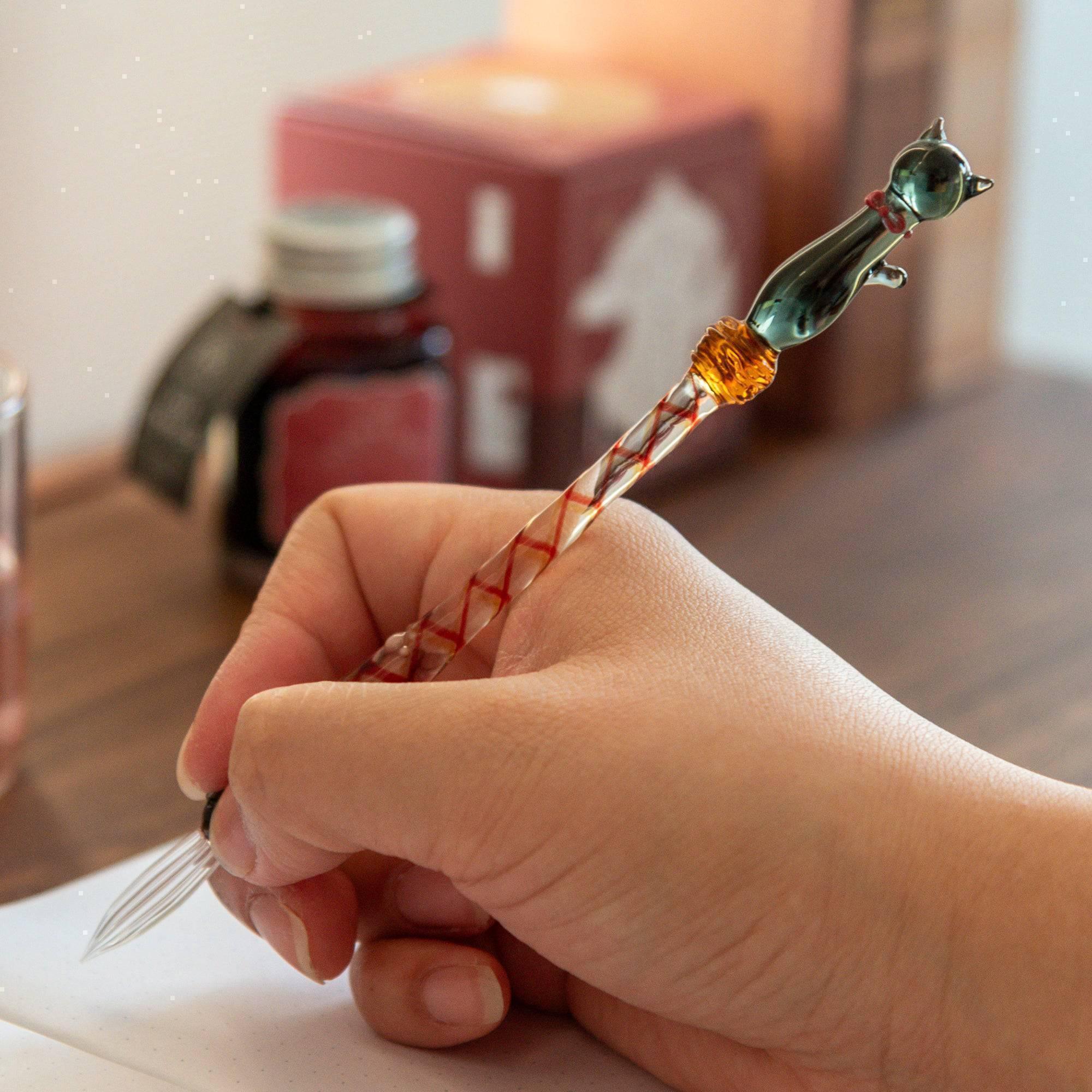 Hand holding a black cat-top glass dip pen with a twisted orange-and-white shaft and a clear glass nib, with an ink bottle blurred in the background.