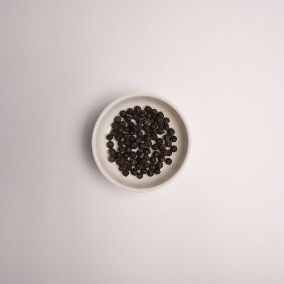 Top-down view of a bowl filled with whole black peppercorns on a plain background.