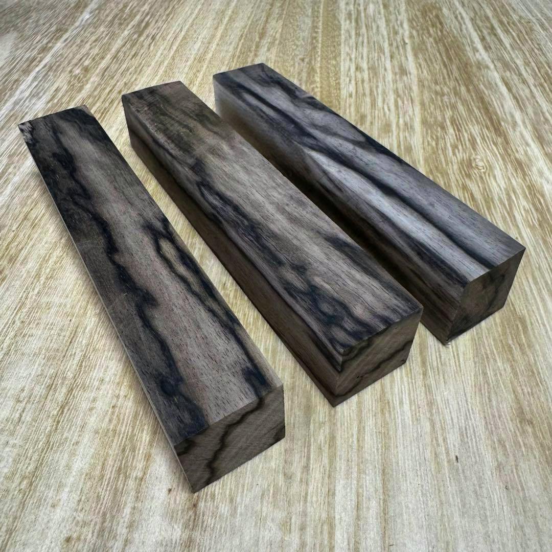 Three black-kaki (black persimmon) pen blanks with peacock grain arranged on a light wood surface.