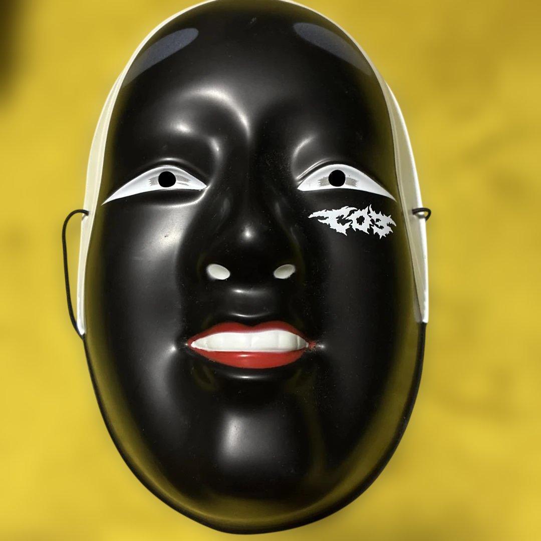 Front view of a black plastic character mask with a smiling face, white eye accents, and red-painted lips against a yellow background.