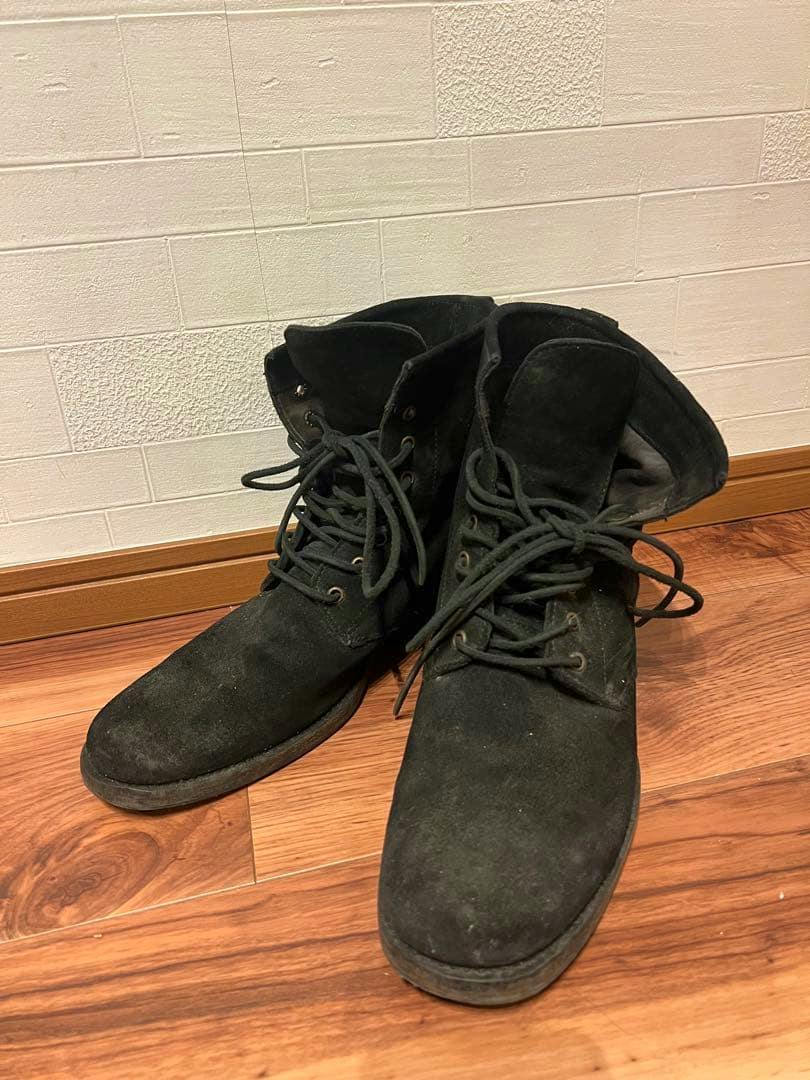 Pair of black suede lace-up boots on a wooden floor against a white textured wall.