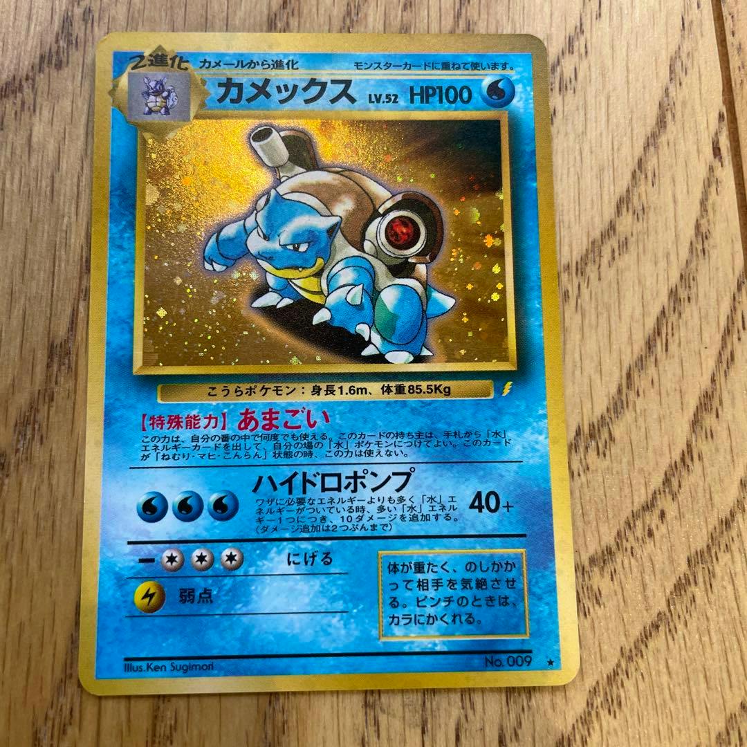 Front of the カメックス (Blastoise) promotional Pokémon trading card showing holographic artwork and Japanese text on a wooden background.