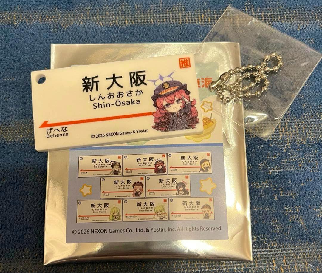 White rectangular Shin-Osaka station-sign style keychain showing “新大阪 / Shin-Osaka” text and a chibi character, shown with its metal ball chain and silver backing packaging.