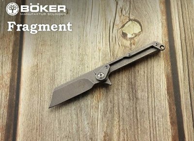 An open BOKER Plus Fragment folding knife displayed on a wooden surface with the BÖKER Fragment logo.