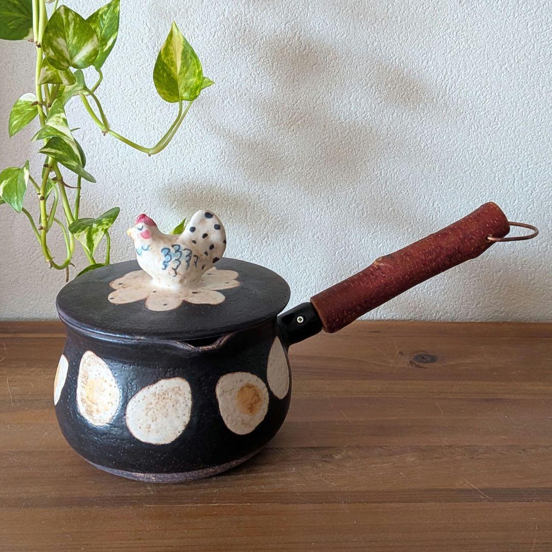A ceramic pot with lid featuring a chicken-shaped knob, black body with white circular patterns and a wooden handle, shown on a wooden surface.