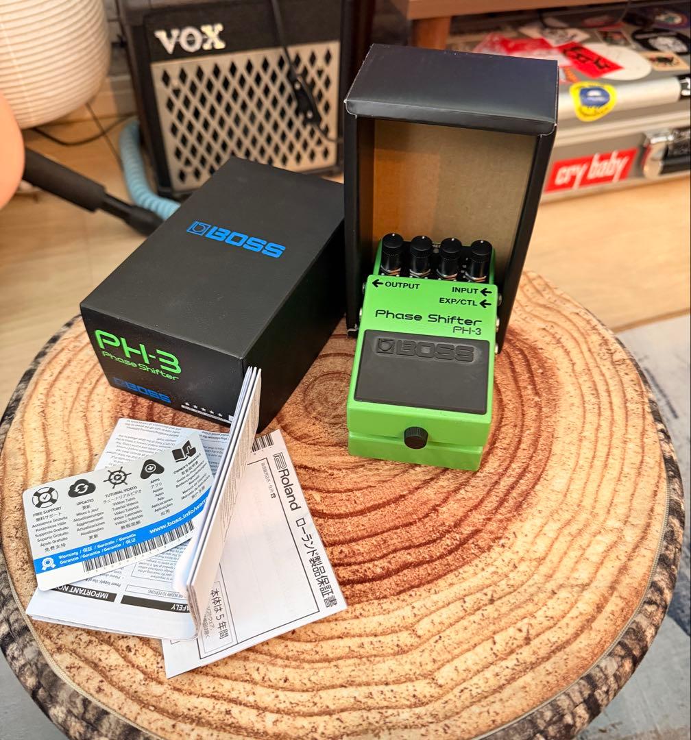 Green BOSS PH-3 Phase Shifter guitar effects pedal displayed next to its box and manuals on a wooden round surface.