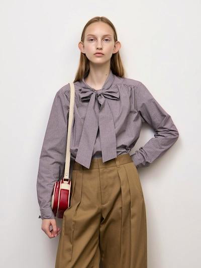 Model wearing a checked bow-tie blouse styled with high-waist brown trousers and a small red shoulder bag.