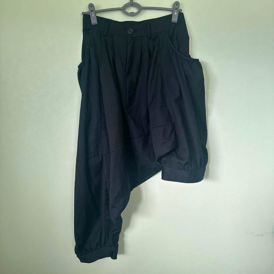 Black asymmetric sarouel women's pants hanging on a hanger against a light wall, showing the front and draped leg.