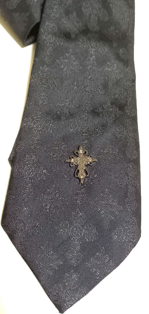 Black patterned polyester necktie photographed flat, showing a small cross-shaped decorative embellishment near the tip.