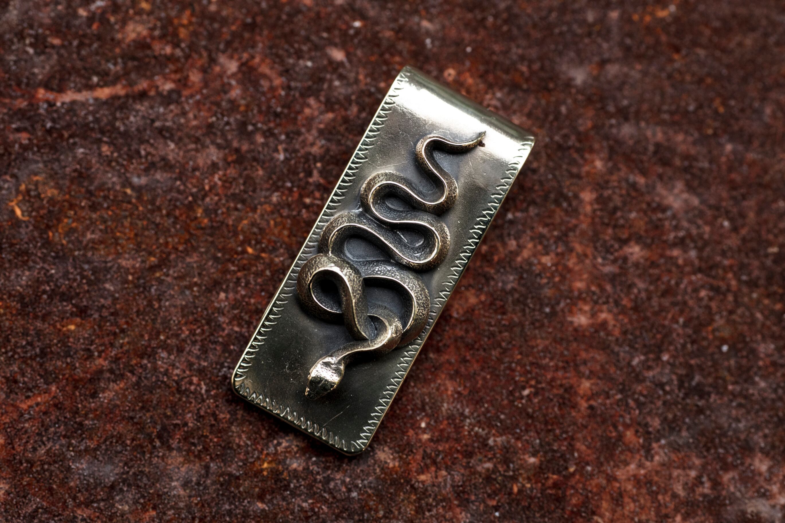 A brass money clip featuring a raised snake design laid on a textured surface.